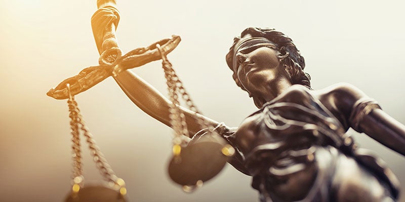 Vista desde un ángulo bajo de una estatua bronceada de la Dama de la Justicia sosteniendo una balanza desequilibrada, con una luz brillante iluminando su rostro.
